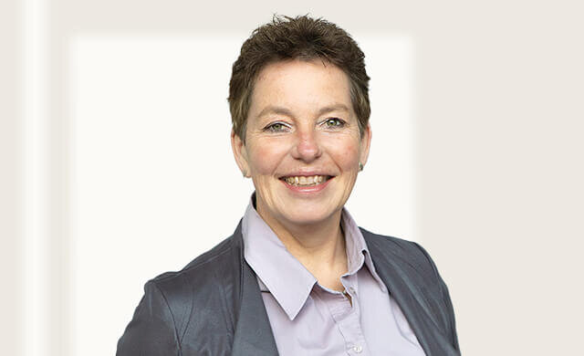 Lächelnde Frau mit kurzem, braunem Haar, trägt eine graue Jacke und ein helles Hemd, vor einem neutralen Hintergrund.