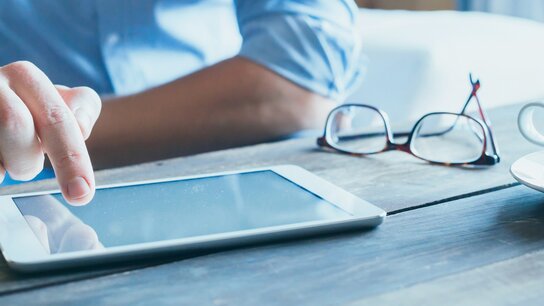 Hand tippt auf ein Tablet, daneben liegen eine Brille und eine Tasse Kaffee auf einem Holztisch.