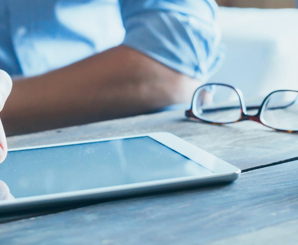 Hand tippt auf ein Tablet, daneben liegen eine Brille und eine Tasse Kaffee auf einem Holztisch.