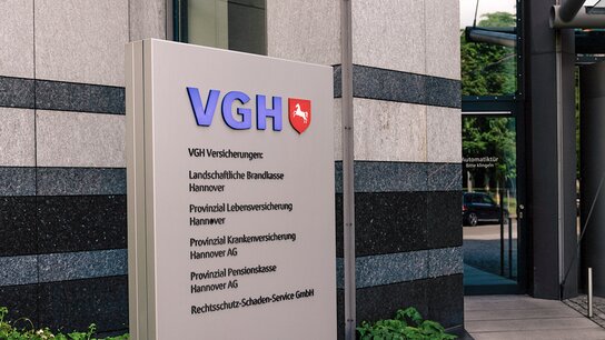 VGH Versicherungsschild sowie Eingangsbereich eines modernen Bürogebäudes mit Drehtür und begrünten Außenanlagen.