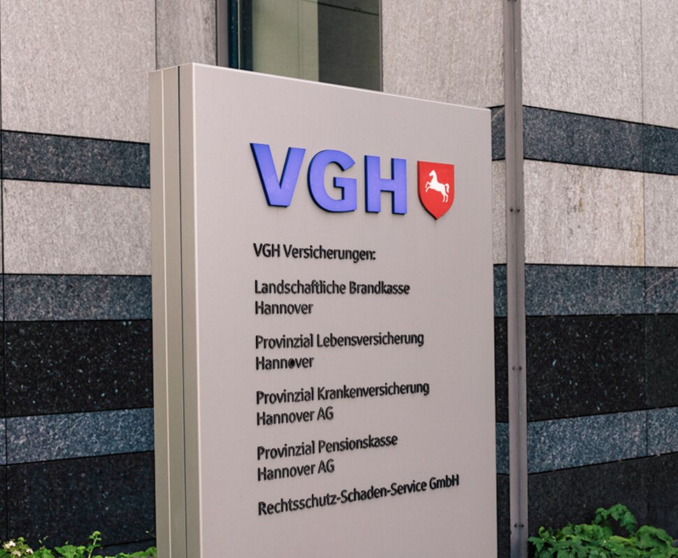 VGH Versicherungsschild sowie Eingangsbereich eines modernen Bürogebäudes mit Drehtür und begrünten Außenanlagen.