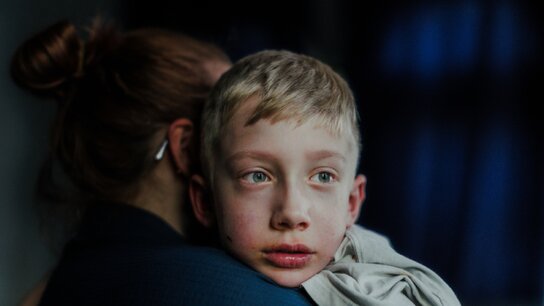 Ein Junge mit müden Augen umschlungen von einer Erwachsenen, im Hintergrund schwaches Licht und Schatten.