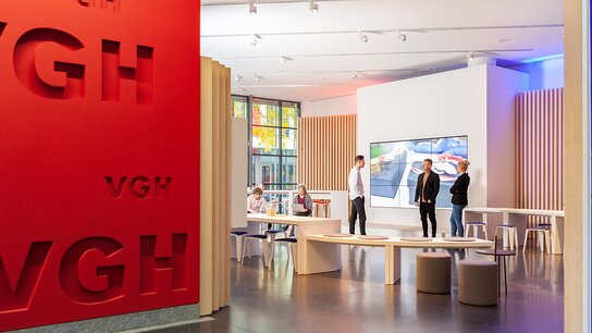 Moderne Ausstellungsräume mit roten und blauen Wänden, interaktiven Displays und mehreren Personen in Diskussion.