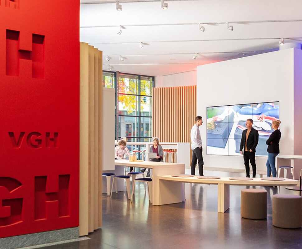 Moderne Ausstellungsräume mit roten und blauen Wänden, interaktiven Displays und mehreren Personen in Diskussion.